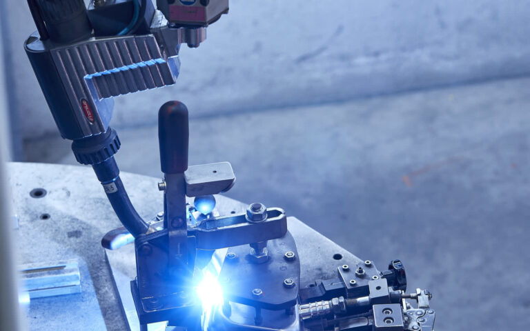 Roboterschweißen mit Fronius-Brenner: blauer Lichtbogen am Metallbauteil in Spannvorrichtung mit Kniehebelspanner