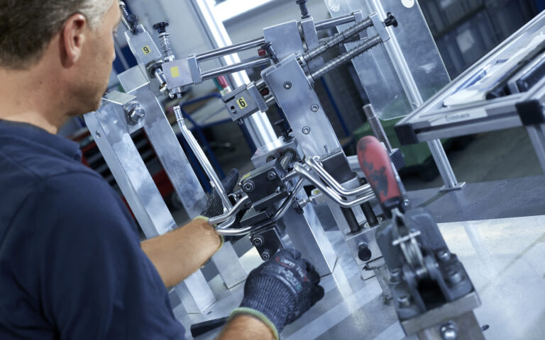 Mitarbeiter richtet gebogenes Metallrohr von Hand in Aluminium-Prüfvorrichtung mit nummerierten Stationen